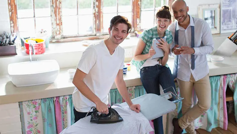 Een man strijkt een shirt terwijl twee mensen toekijken. Een wastafel en ramen zijn op de achtergrond te zien.