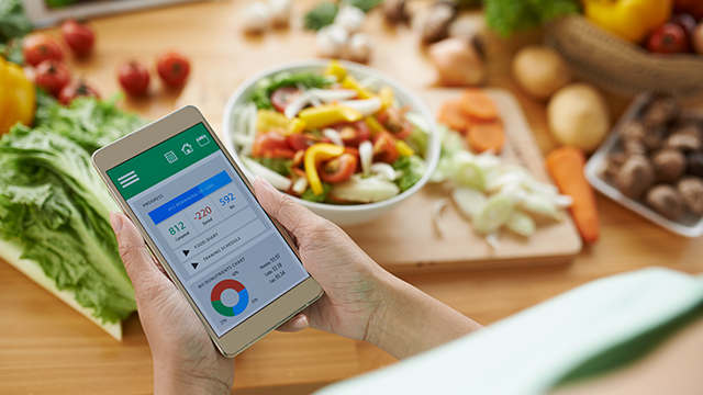 Een persoon houdt een telefoon vast met een app die voedingstracking weergeeft, naast een salade en verse groenten op een houten tafel.