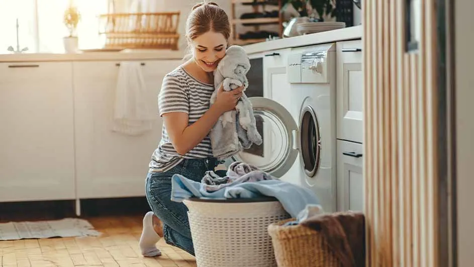 Vrouw bij wasmachine, met wasmand vol kleding. Ze houdt een stuk wasgoed vast. Keukenachtergrond.