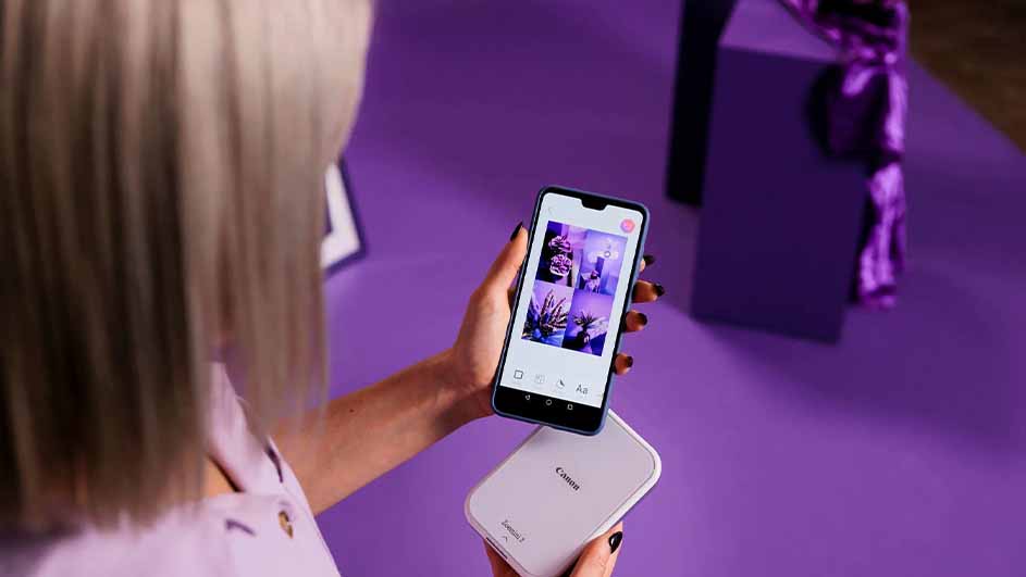 Een vrouw houdt een telefoon met foto-app boven een witte Canon printer op een paarse achtergrond.