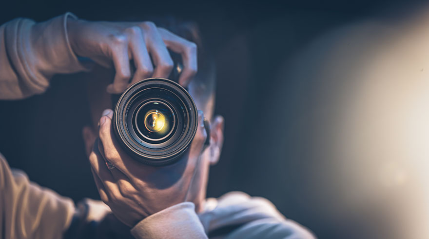 Een persoon houdt een zwarte cameralens vast, met een lichtbron zichtbaar in het midden van de lens. De achtergrond is donker.