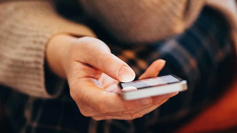Een hand houdt een witte smartphone vast, de vinger raakt het scherm aan, op de achtergrond is een beige trui te zien.