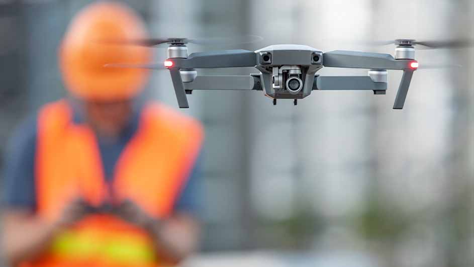 Een grijze drone vliegt; op de achtergrond een persoon met een oranje veiligheidshelm en vest.