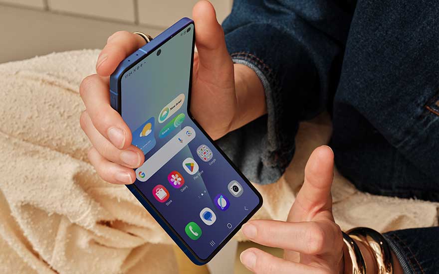 A hand holds a blue foldable smartphone half open, with apps visible on the screen and soft fabrics in the background.