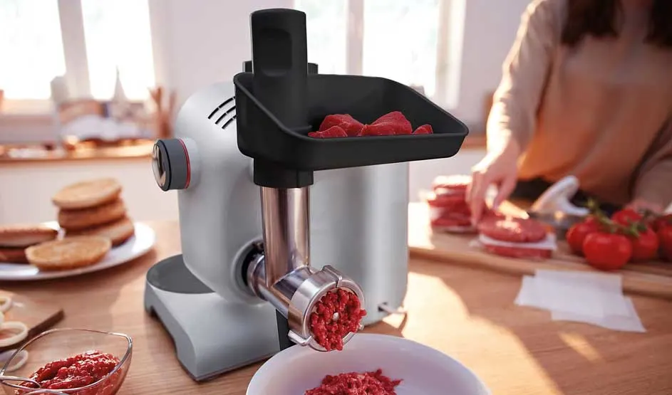 Een vleesmolen op het aanrecht maalt rood vlees in een kom; achteraan snijdt iemand burgers, met broodjes en tomaten in warm licht.