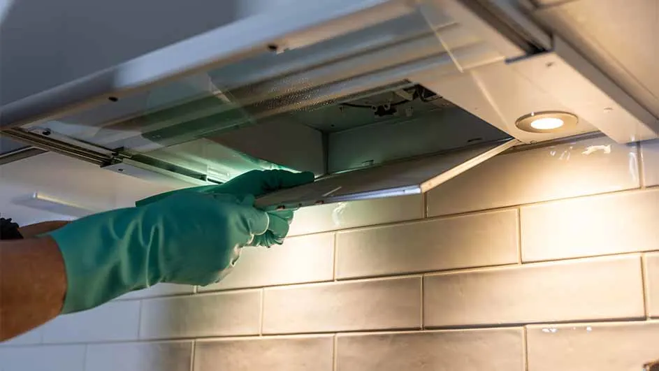 A hand in a green glove opens a range hood filter. The hood has a light. The background is a tiled wall.