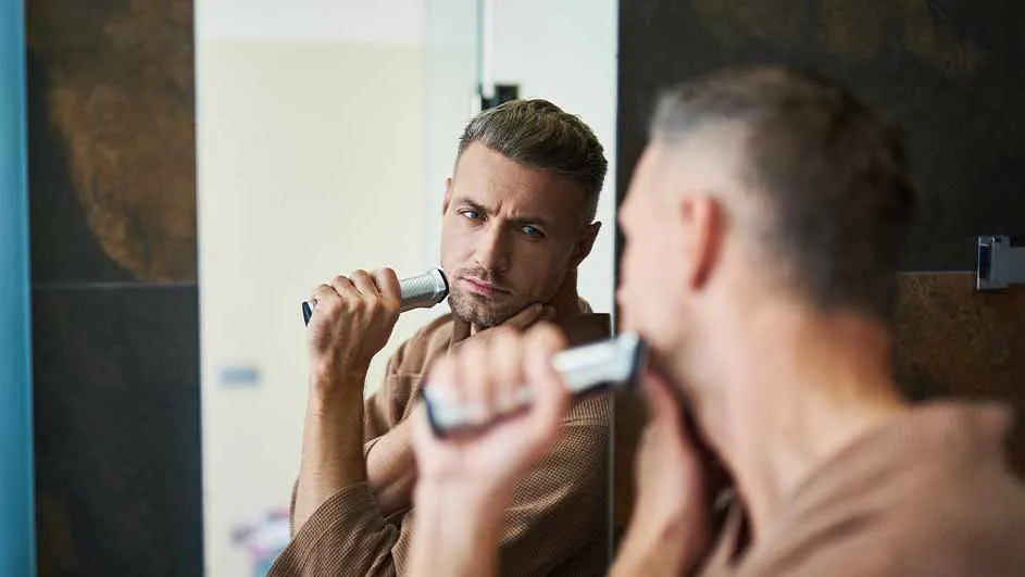 Een man in een bruine badjas scheert zich voor een spiegel met een zilverkleurig elektrisch scheerapparaat in een badkamer.