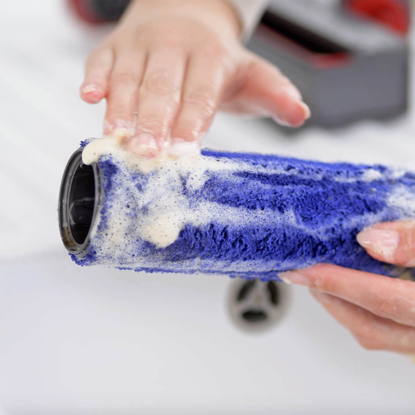 Handen wassen een blauwe borstel met zeep boven een witte gootsteen. Op de achtergrond is een stofzuiger te zien.