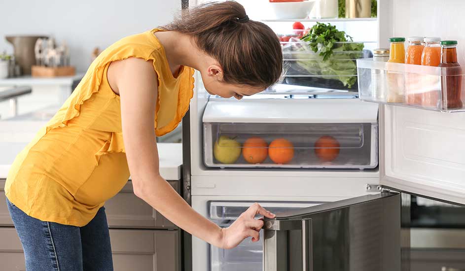 Een vrouw in een gele top opent een koelkast vol groenten, fruit en sapjes. Het merk van de koelkast is niet zichtbaar.