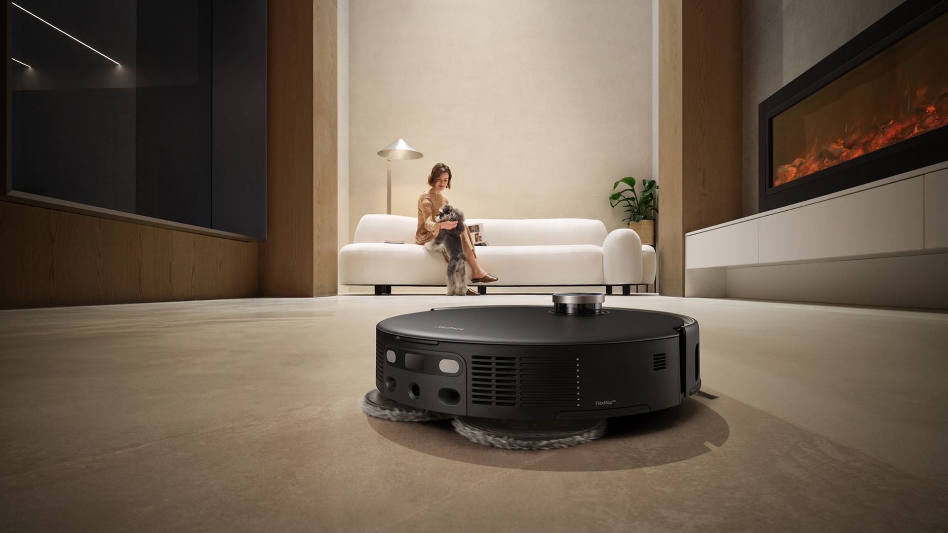 Een zwarte robotstofzuiger maakt een kamer schoon. Een vrouw zit op een witte bank met een hond. Een open haard brandt. De kamer is modern.