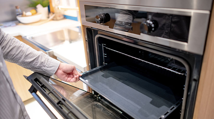 Een hand opent de deur van een roestvrijstalen oven in een keuken, met een bakplaat zichtbaar.