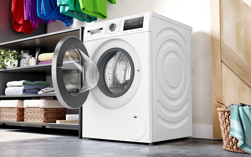 A white Bosch washing machine with open door stands in a laundry area. Colourful clothes (red, purple, blue, green) hang above. To the left is a black shelving unit with folded towels and baskets. To the right, a wicker basket is visible next to a wooden wall.