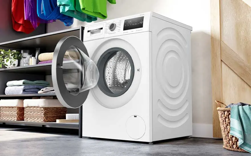 A white Bosch washing machine with open door stands in a laundry area. Colourful clothes (red, purple, blue, green) hang above. To the left is a black shelving unit with folded towels and baskets. To the right, a wicker basket is visible next to a wooden wall.