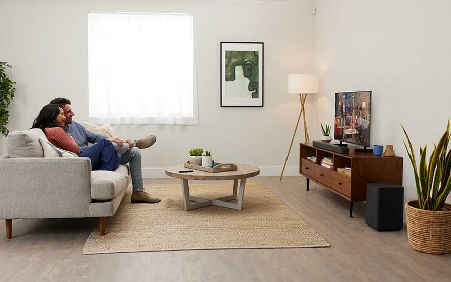 Stel zit op lichte bank in minimalistische woonkamer met kleine tv, houten meubel, planten, lamp en vloerkleed.