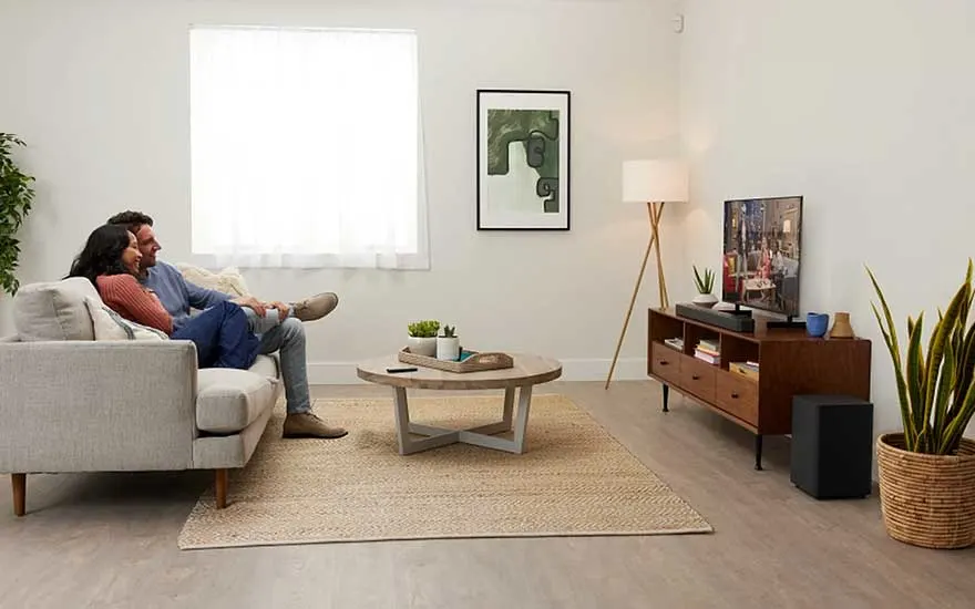 Stel zit op lichte bank in minimalistische woonkamer met kleine tv, houten meubel, planten, lamp en vloerkleed.