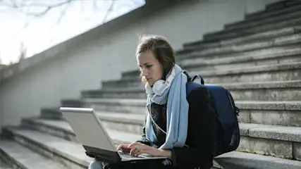 Een vrouw zit op een stenen trap met een laptop. Ze draagt een blauwe sjaal, een rugzak en een koptelefoon om haar nek.