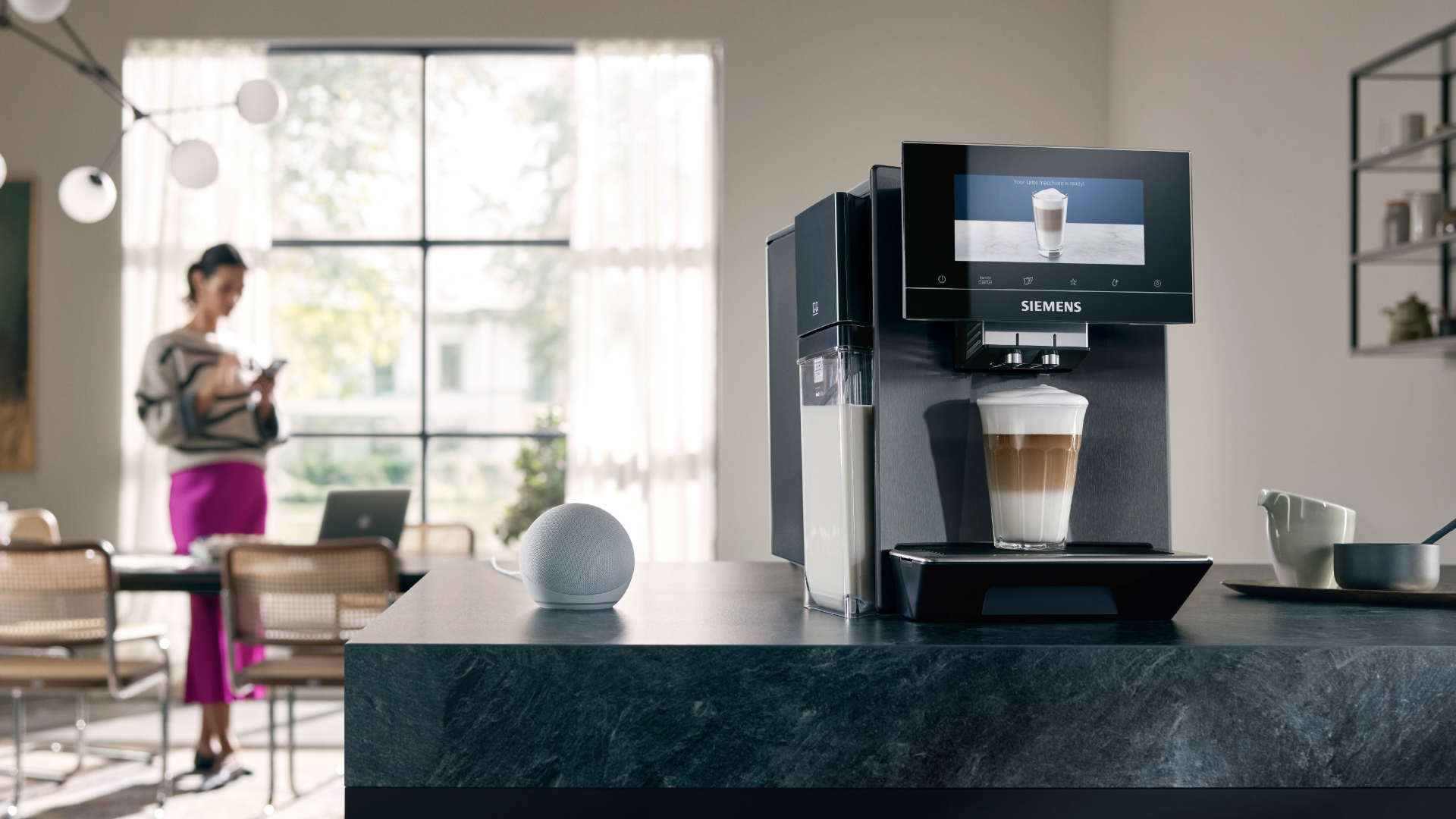 Een koffiemachine maakt een latte macchiato in een keuken. Een vrouw gebruikt een telefoon op de achtergrond.