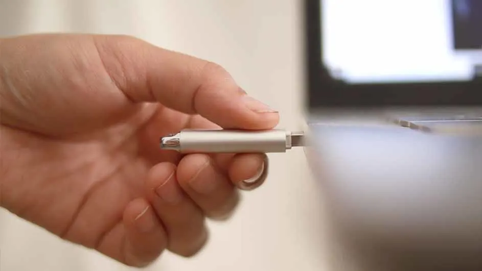 Een hand steekt een zilverkleurige USB-stick in een laptop. De achtergrond is wazig en licht.