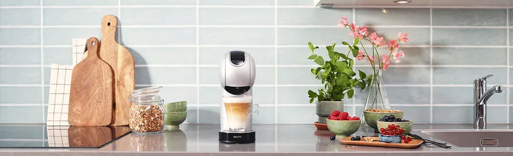Op het aanrecht staat een zilveren Krups-koffiemachine met een glas cappuccino, omringd door fruit, granola en groene planten.