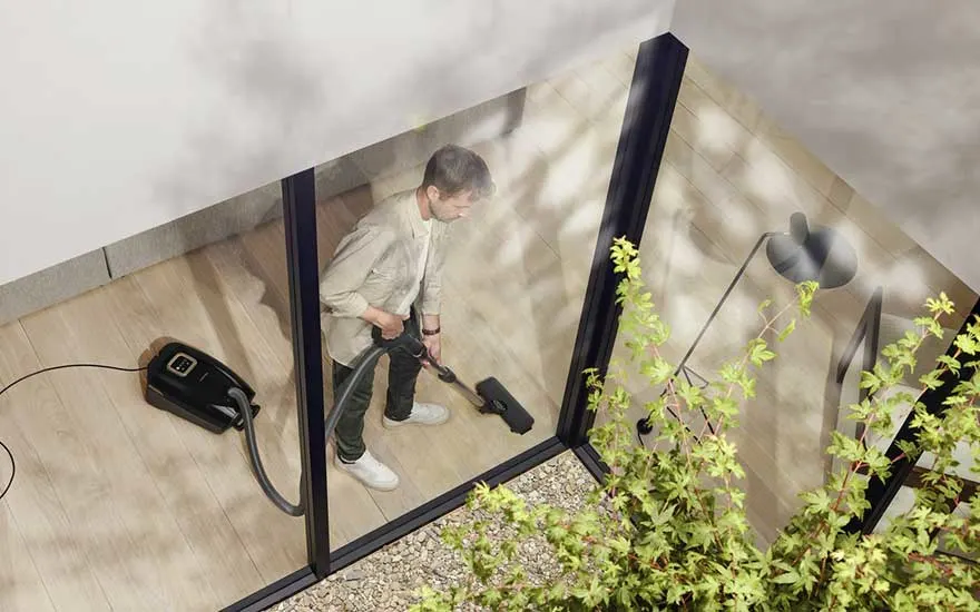 Een man stofzuigt de vloer terwijl hij zichtbaar is door het raam, in een moderne woning met houten vloer en grote ramen.