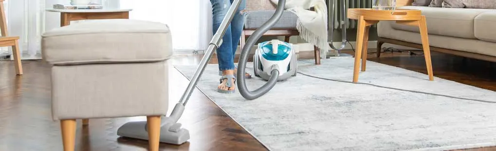A person hoovers a pale rug in a bright living room, with wooden furniture, soft textiles and a compact cylinder vacuum cleaner.