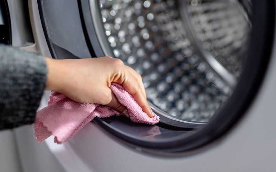 Een persoon maakt de rubberen rand van een wasmachinedeur schoon met een roze microvezeldoek. De trommel van de wasmachine is vaag zichtbaar achter de rand, en het geheel is goed verlicht.