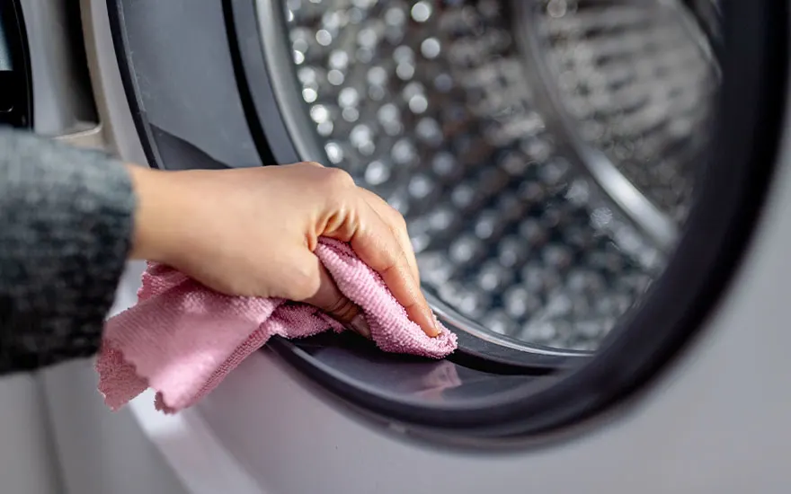 A person is cleaning the rubber seal around a washing machine door with a pink microfibre cloth. The washing machine drum is visible in the background, and the scene is well-lit.