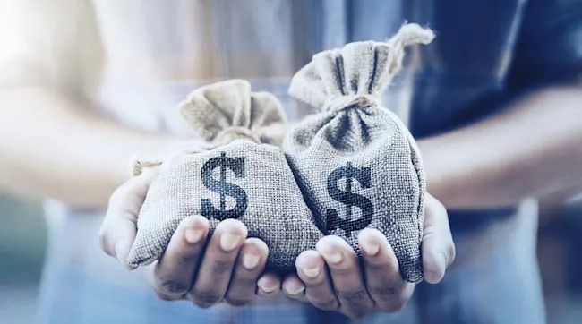 Twee handen houden twee zakken vast met een dollarteken erop, tegen een vage, blauwe achtergrond.