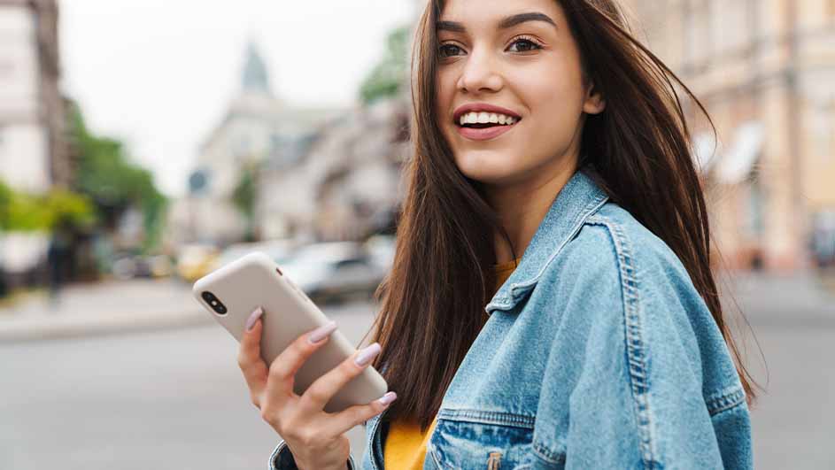 Jonge vrouw met een telefoon in de hand, gekleed in een spijkerjas, op straat met gebouwen op de achtergrond.