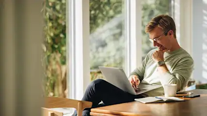 Man zit aan een houten tafel met een laptop, kopje, notitieboek en telefoon. Achter hem een groot raam met groen uitzicht.