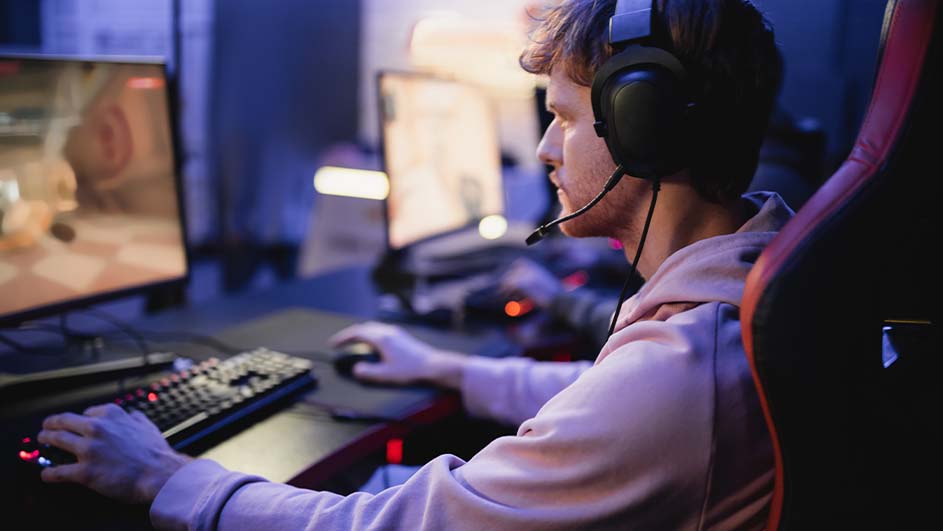 Een persoon met een headset speelt een computerspel. Er zijn schermen, een toetsenbord en een muis zichtbaar in de blauwe kamer.