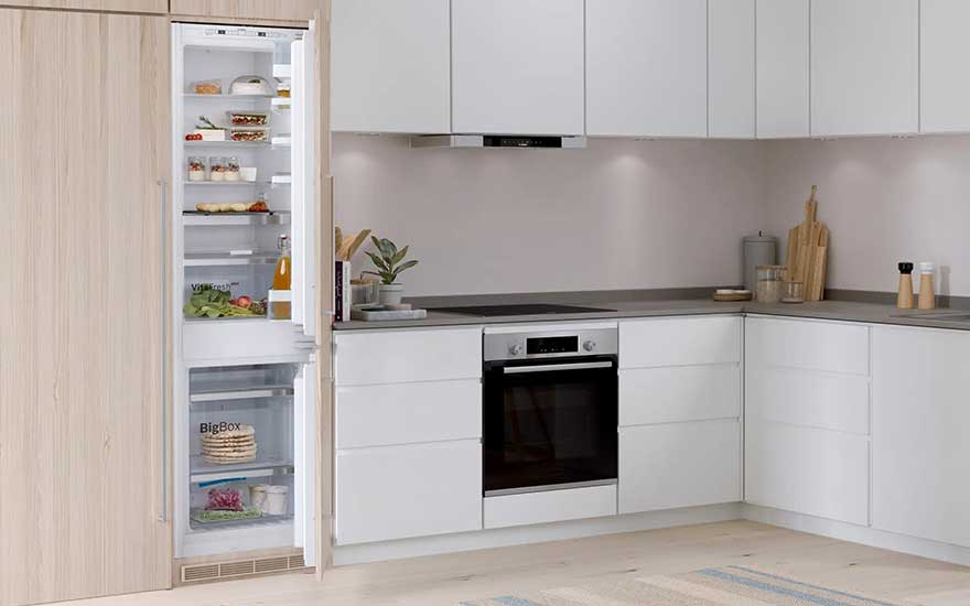 A Bosch fridge, filled with food, stands open in a sleek white kitchen with modern cabinets, dark worktops, and built-in appliances.