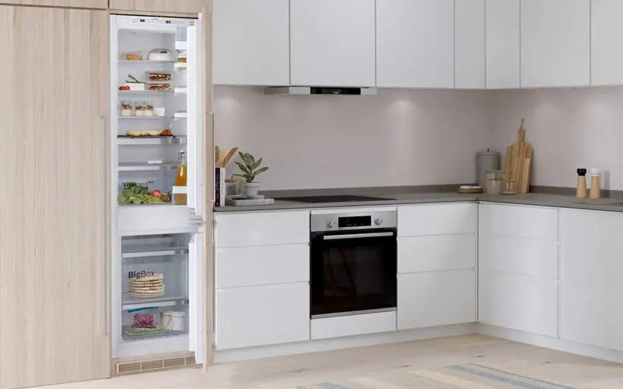 A Bosch fridge, filled with food, stands open in a sleek white kitchen with modern cabinets, dark worktops, and built-in appliances.