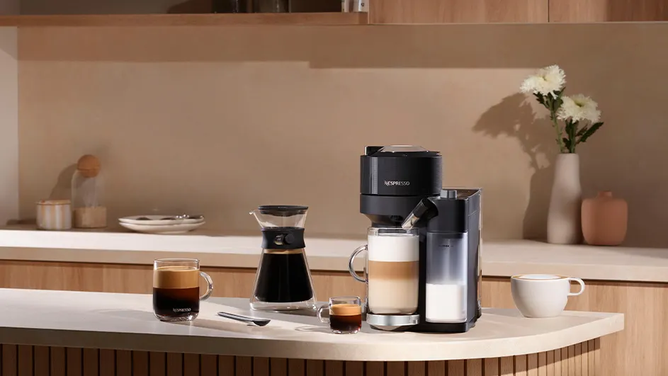 Een aanrecht met koffiezetapparaten, glazen koffie, een theelepel en een kopje tegen een beige achtergrond.