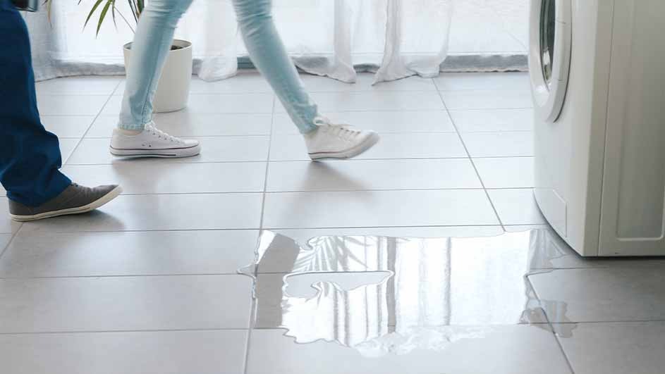 Tiled floor with water puddle, near a washing machine. Two people's feet are visible, one in blue pants and another in light pants.