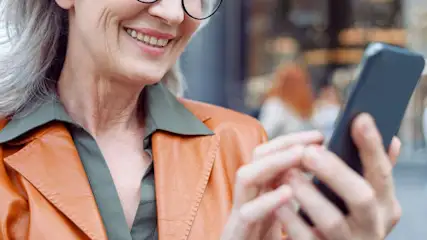 A smiling woman wearing glasses and an orange jacket is outdoors, holding and using a smartphone. No visible brand appears on the phone or clothing.