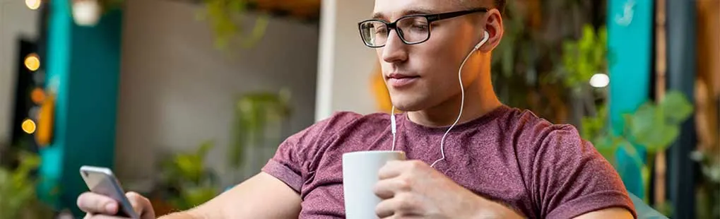 Jonge man met bril zit ontspannen in een café, luistert met bedrade oordopjes naar zijn telefoon terwijl hij een warme mok vasthoudt.