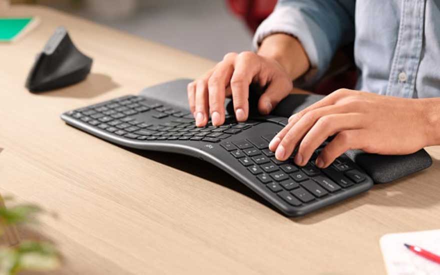 Iemand typt op een ergonomisch, gebogen toetsenbord met polssteun aan een houten bureau.