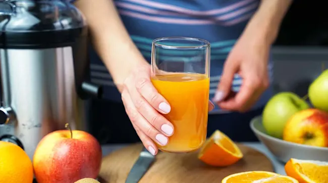 Een persoon houdt een glas sinaasappelsap vast, met een sapcentrifuge en fruit op de achtergrond.