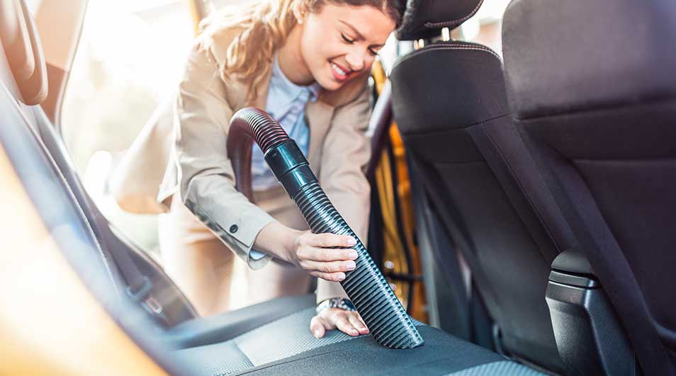 A woman is attentively vacuuming the inside of her car, focusing on cleaning the back seat to keep everything tidy and spotless.