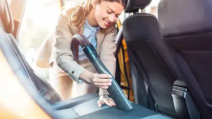 A woman is attentively vacuuming the inside of her car, focusing on cleaning the back seat to keep everything tidy and spotless.