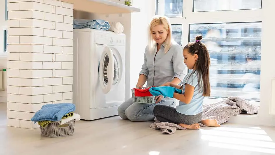 Moeder en dochter knielen bij een wasmachine, met wasmanden en kleding in een lichte ruimte.