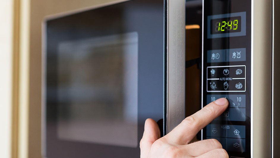 Een vinger drukt op de knop van een magnetron met een digitaal display dat "12:49" aangeeft.