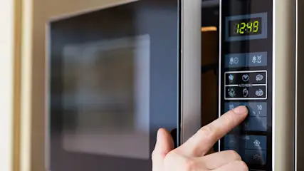 Een vinger drukt op de knop van een magnetron met een digitaal display dat "12:49" aangeeft.