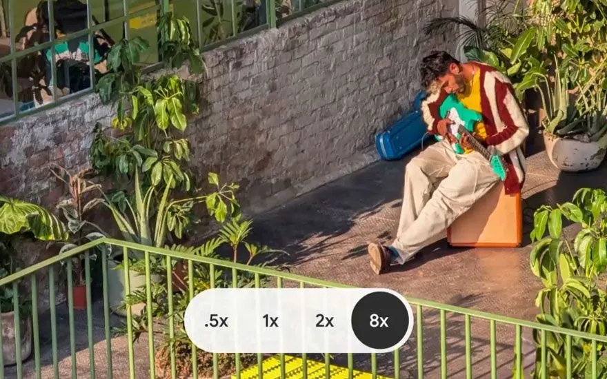 Smartphone camera-interface toont zoom opties (.5x tot 8x) terwijl iemand op een terras zit. Demonstratie van geavanceerde zoom functionaliteit.