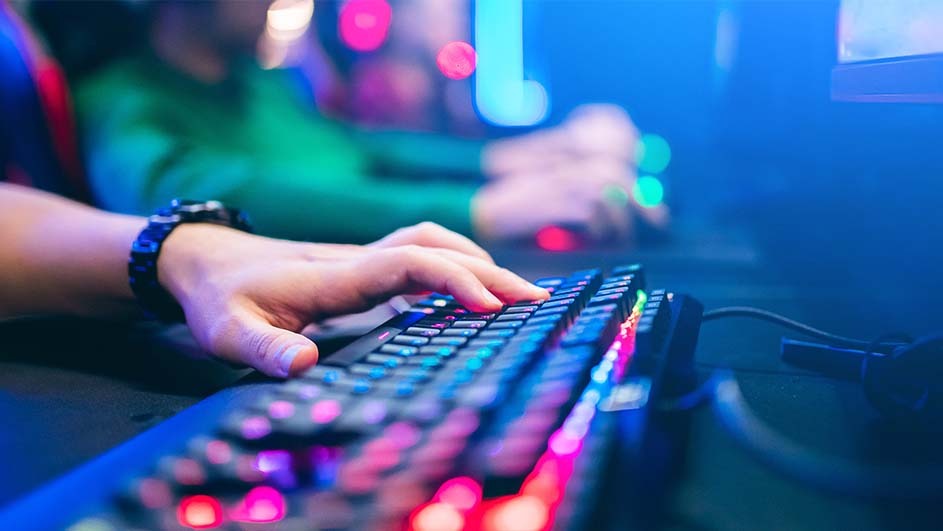 A hand with a watch types on a lit keyboard. Another person is in the background. Blue light is present.
