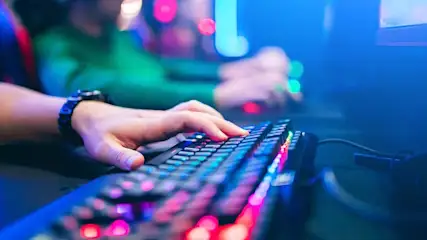 A hand with a watch types on a lit keyboard. Another person is in the background. Blue light is present.