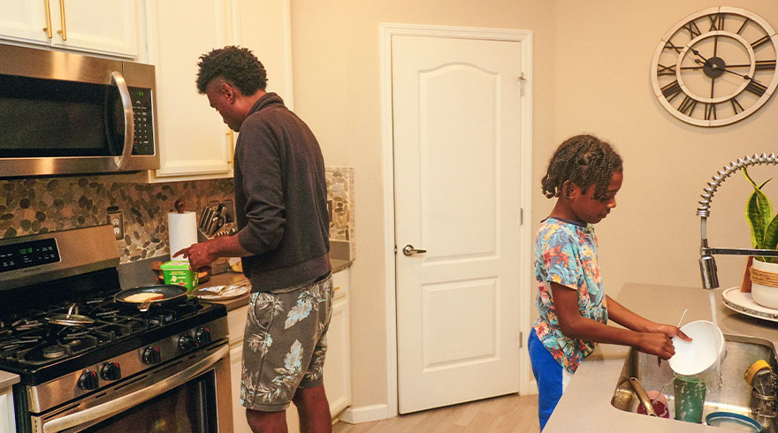 Een man kookt bij een fornuis, terwijl een kind afwast in een keuken met witte kasten en een grote klok.