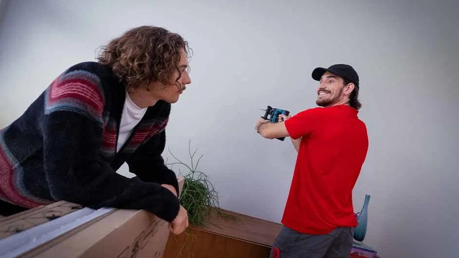 Man met krullend haar leunt op een doos, terwijl een man in een rood shirt een nietpistool vasthoudt tegen een witte achtergrond.