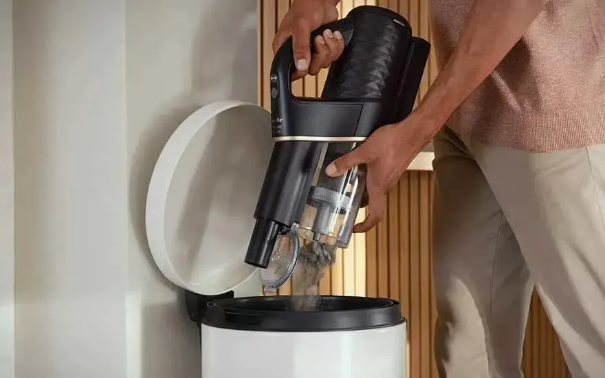 A person empties the dust container of a black stick vacuum cleaner into a bin, with the lid open, in a modern room.